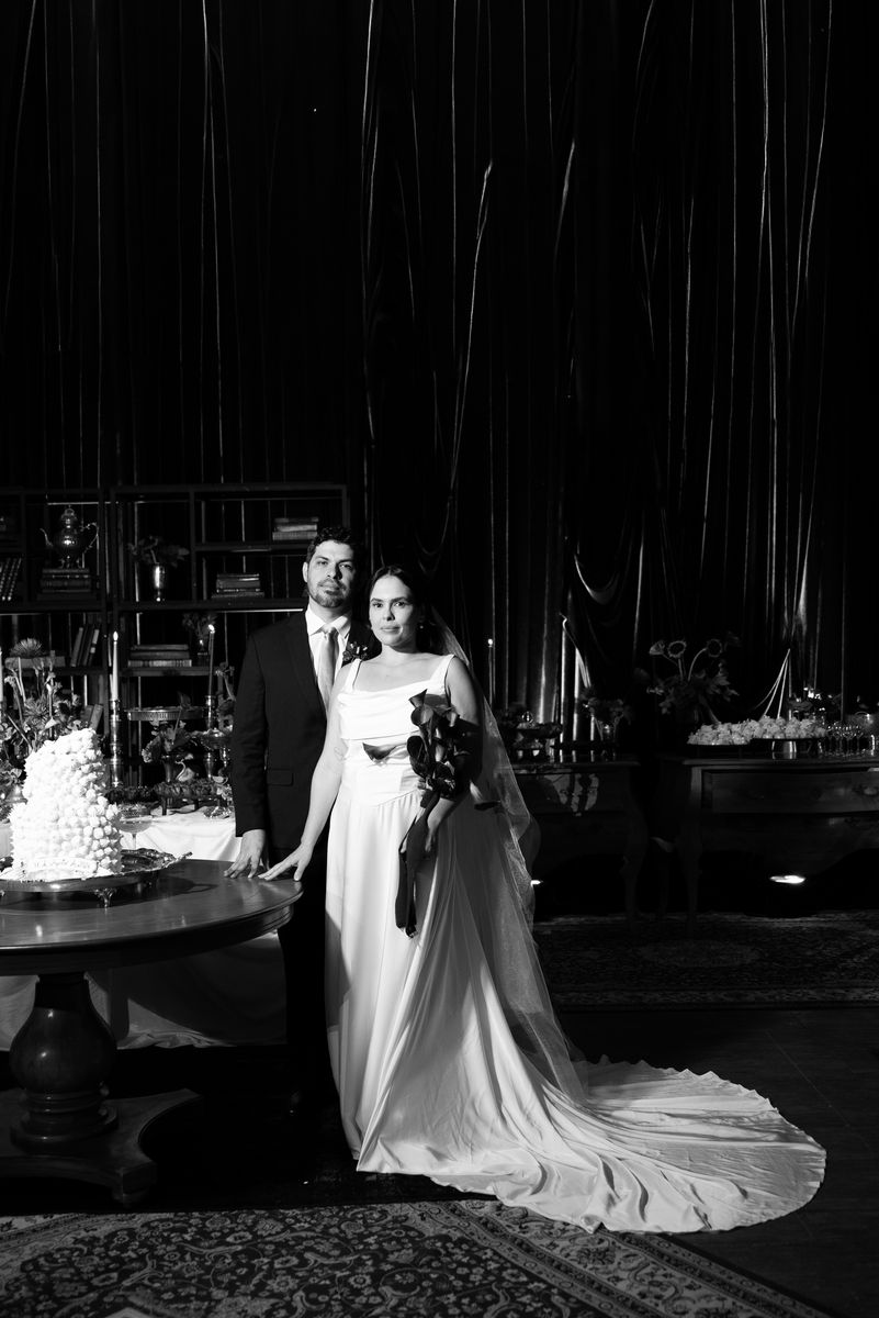 Noivos posando ao lado do bolo, em preto e branco